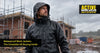 Tradesperson in a hooded waterproof work jacket with rain beading on the fabric, standing on a wet UK construction site with scaffolding behind